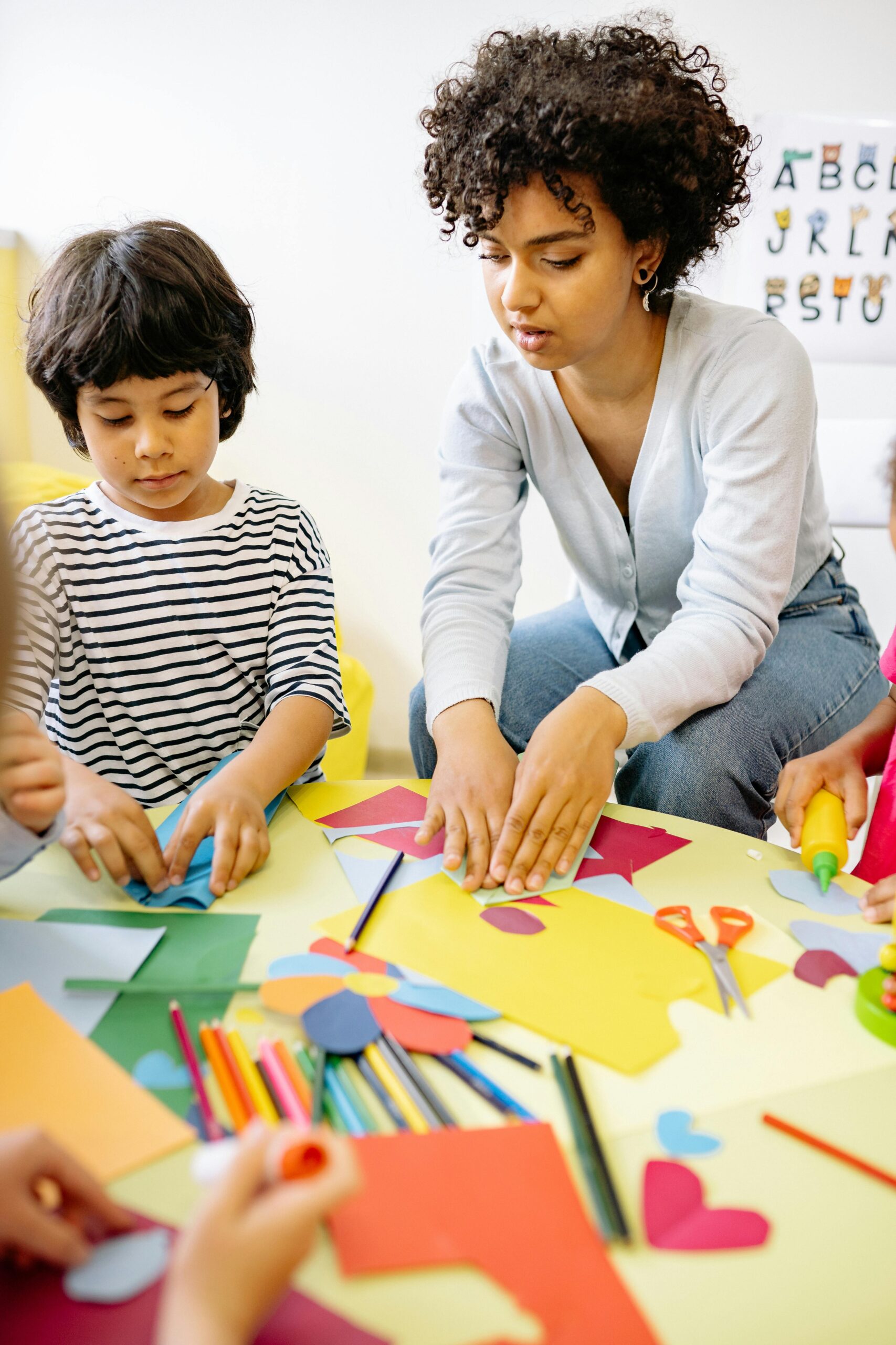 A teacher and young children sit at a table doing arts and crafts with colorful paper, pencils, scissors, and glue.
